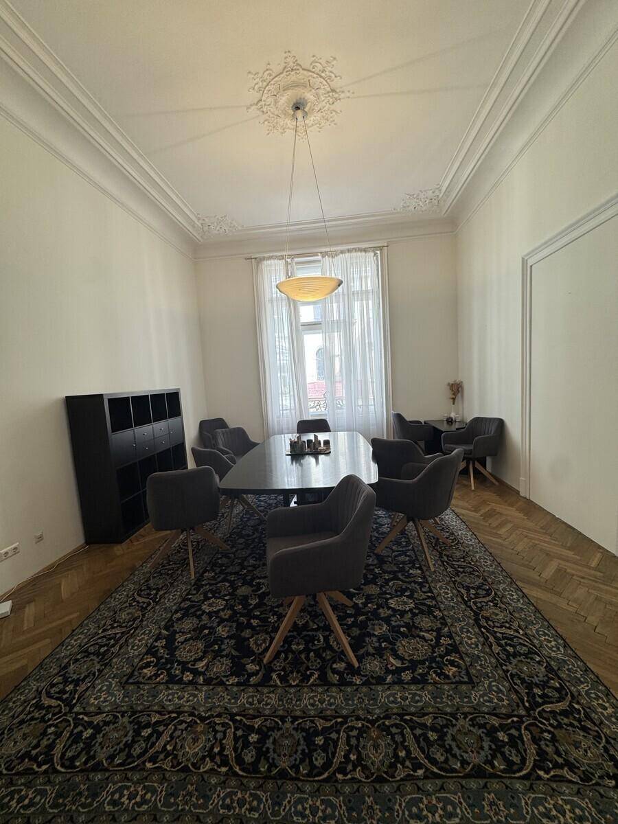 Esszimmer mit großem Tisch, Stühlen, Fischgrätparkett und einem gemusterten Teppich unter einem Deckenleuchter.