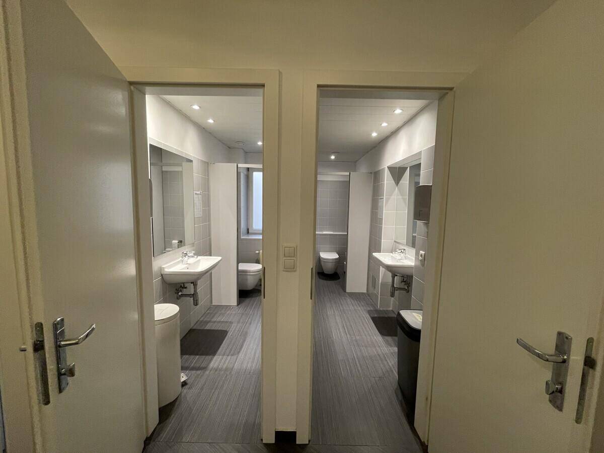 Zwei moderne Badezimmer mit Waschbecken, Toiletten und hellen Fliesen, durch offene Türen sichtbar.