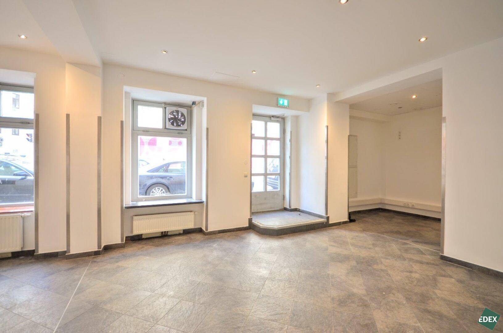 Heller Geschäftsraum mit Fliesenboden, großen Fenstern und einem sichtbaren Abluftventilator im Fensterbereich.