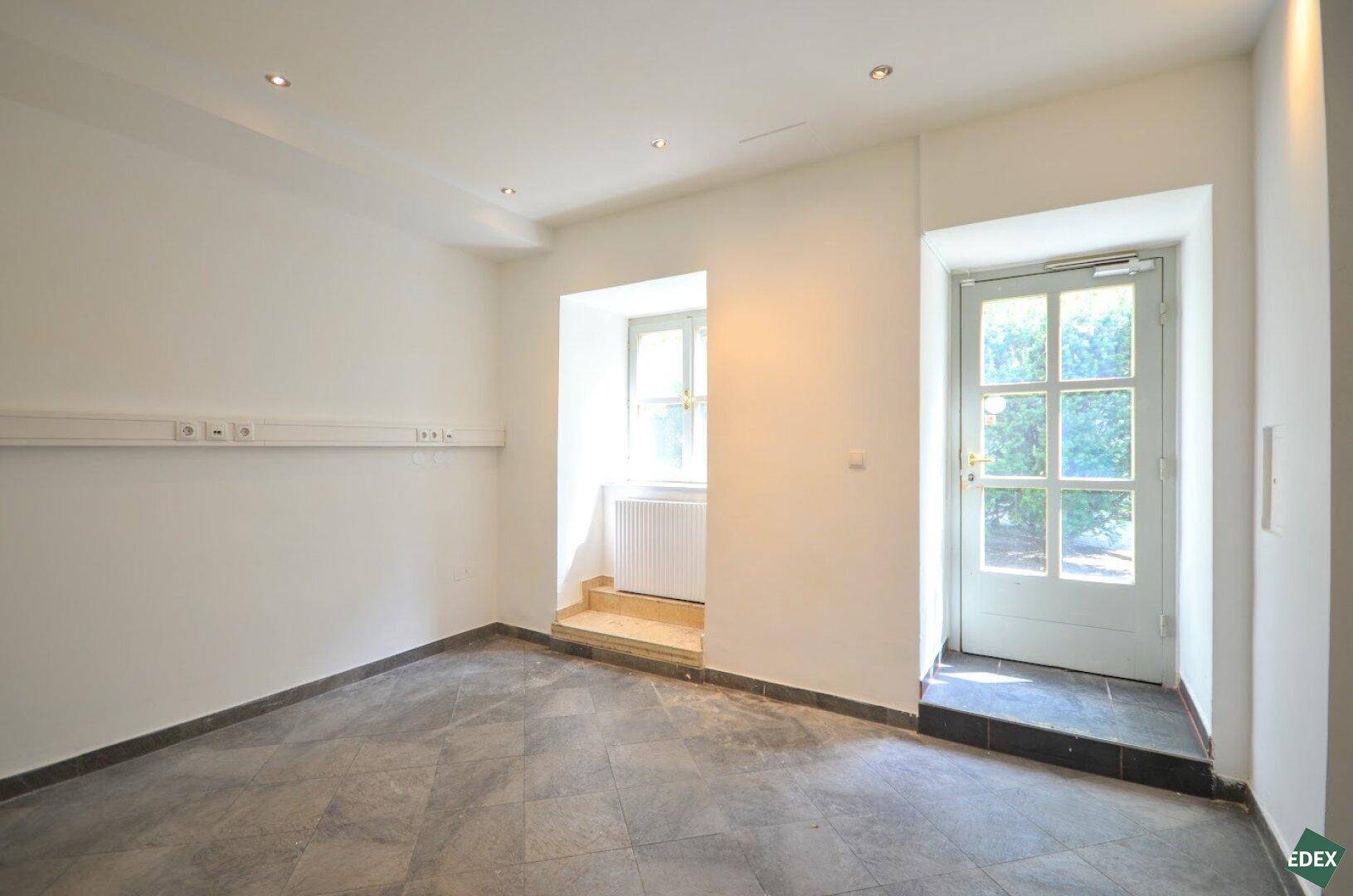 Heller, leerer Raum mit Fliesenboden, einem Fenster und einer Eingangstür mit Glaspaneelen zur Straße.