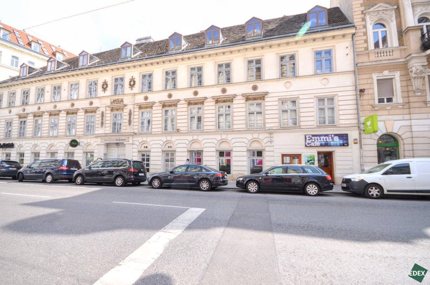 Historisches Gebäude mit klassischer Fassade, vielen Fenstern und Geschäften im Erdgeschoss an einer belebten Straße.