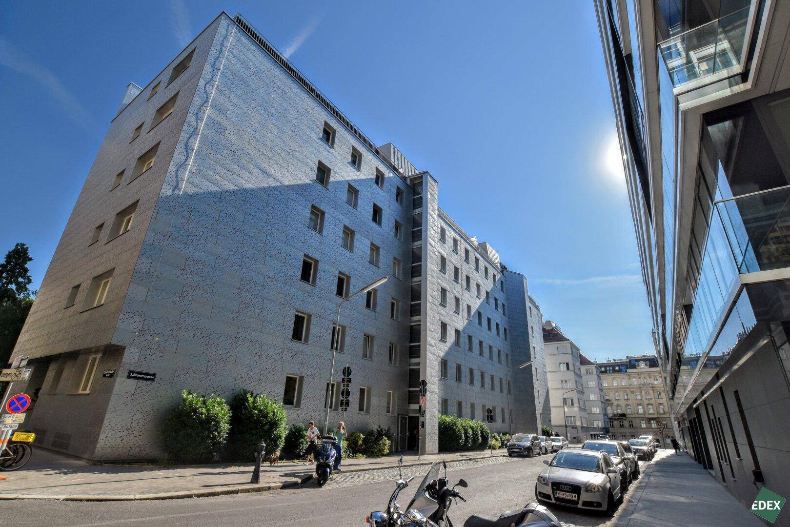 Modernes Mehrfamilienhaus an einer belebten Straße unter blauem Himmel, mit vielen Fenstern.