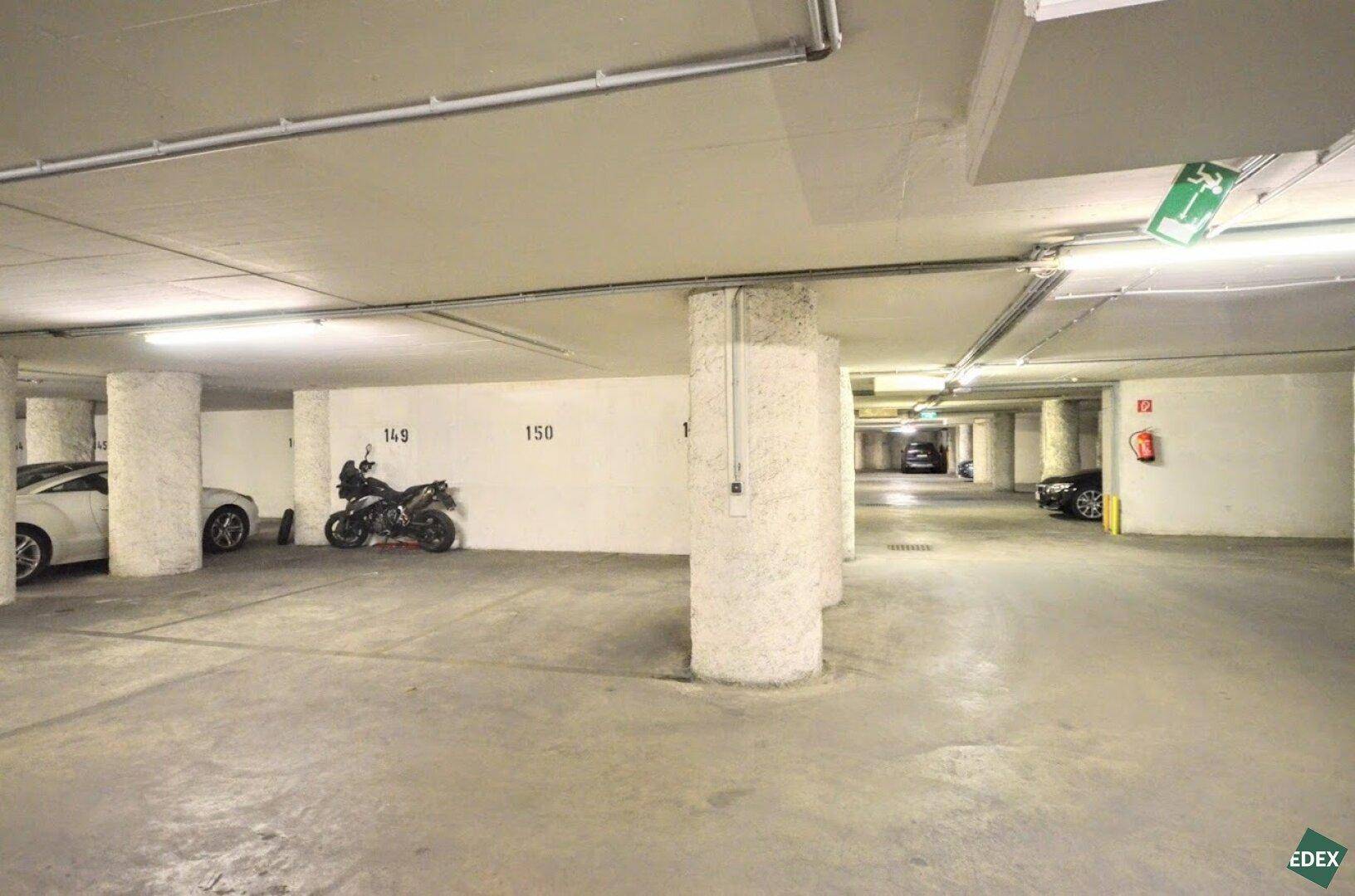 Helle Tiefgarage mit mehreren Fahrzeugen, Betonstützen und einem sichtbaren Notausgangsschild an der Decke.