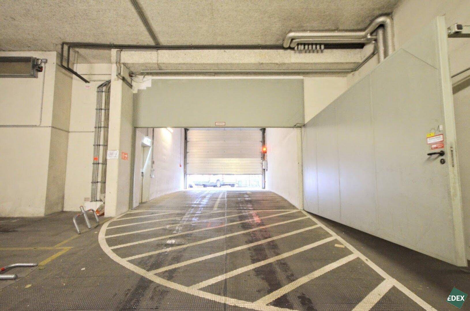 Einfahrt zu einer Tiefgarage mit Rampe und Rolltor, die gut beleuchtet ist.