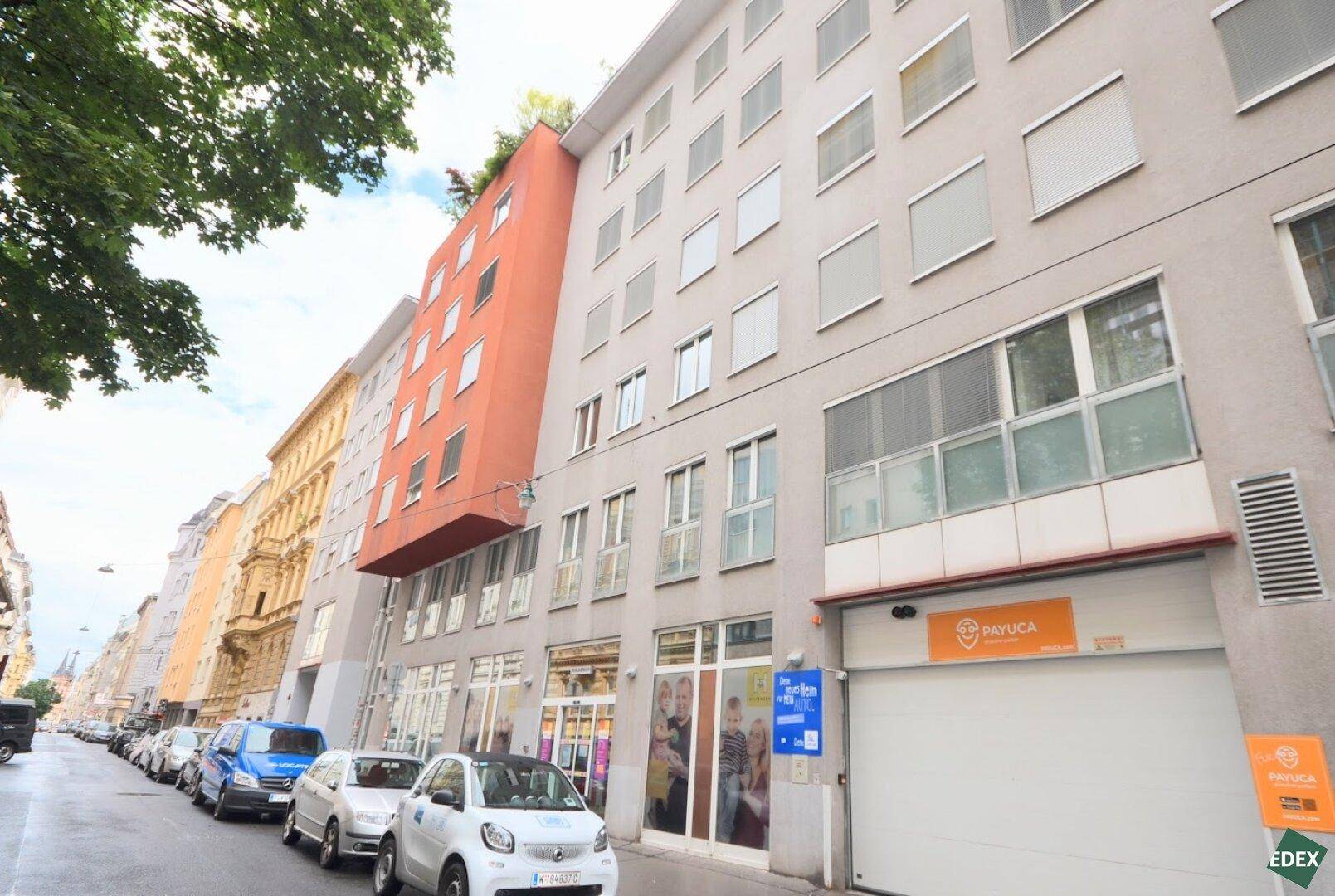 Ansicht eines modernen Mehrfamilienhauses mit Geschäften im Erdgeschoss und geparkten Autos auf der Straße.
