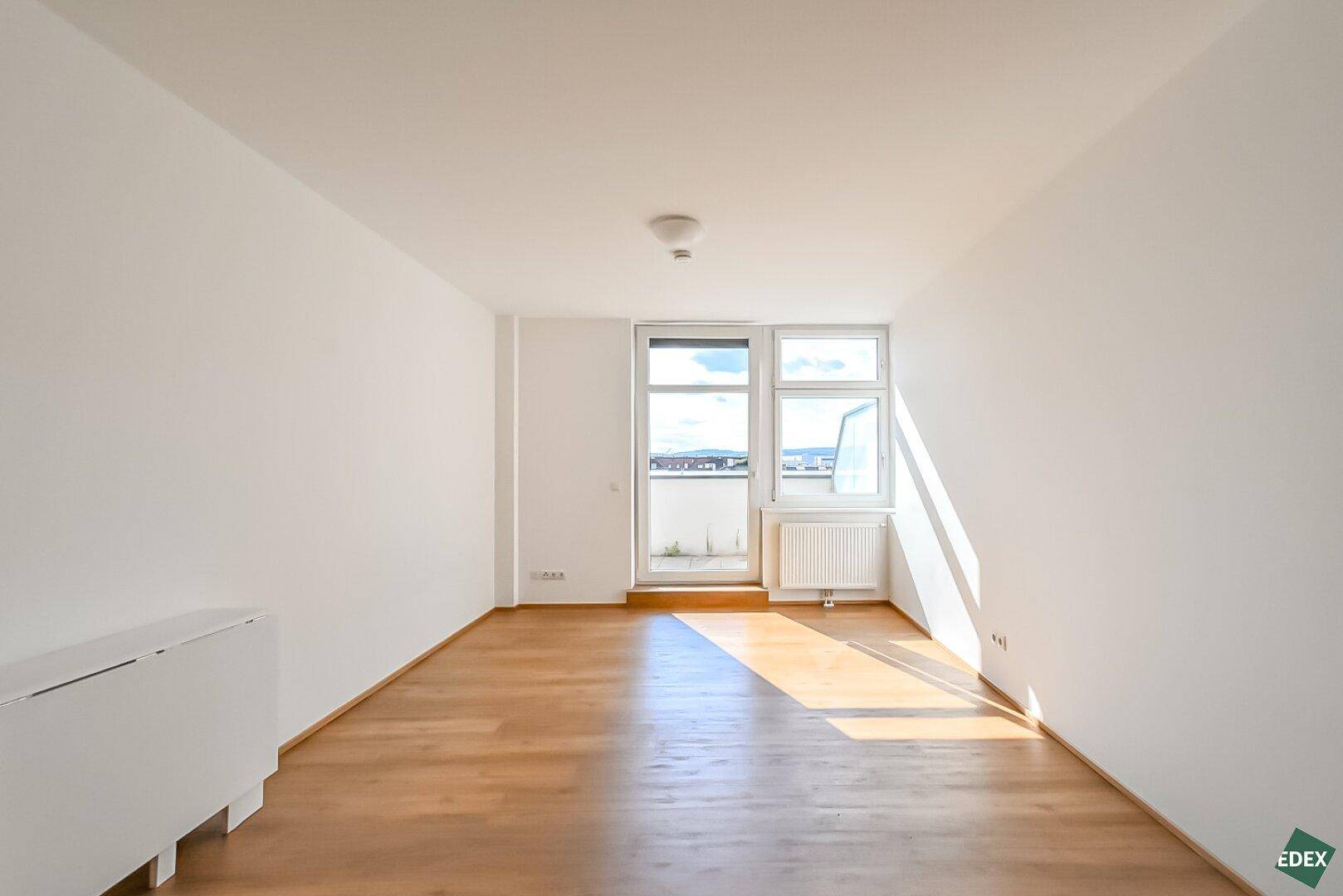 Heller, leerer Wohnraum mit Holzboden, großen Fenstern und Zugang zu einem Balkon.