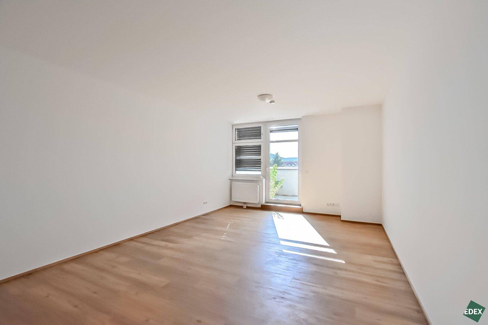 Geräumiger, heller Wohnbereich mit Holzboden und Fensterfront, die viel Tageslicht hereinlässt.