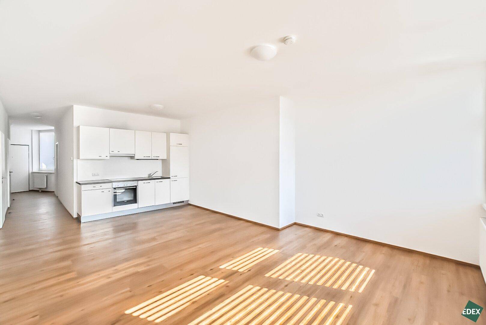 Heller Wohn- und Küchenbereich mit weißer Einbauküche, Holzboden und viel Tageslicht.