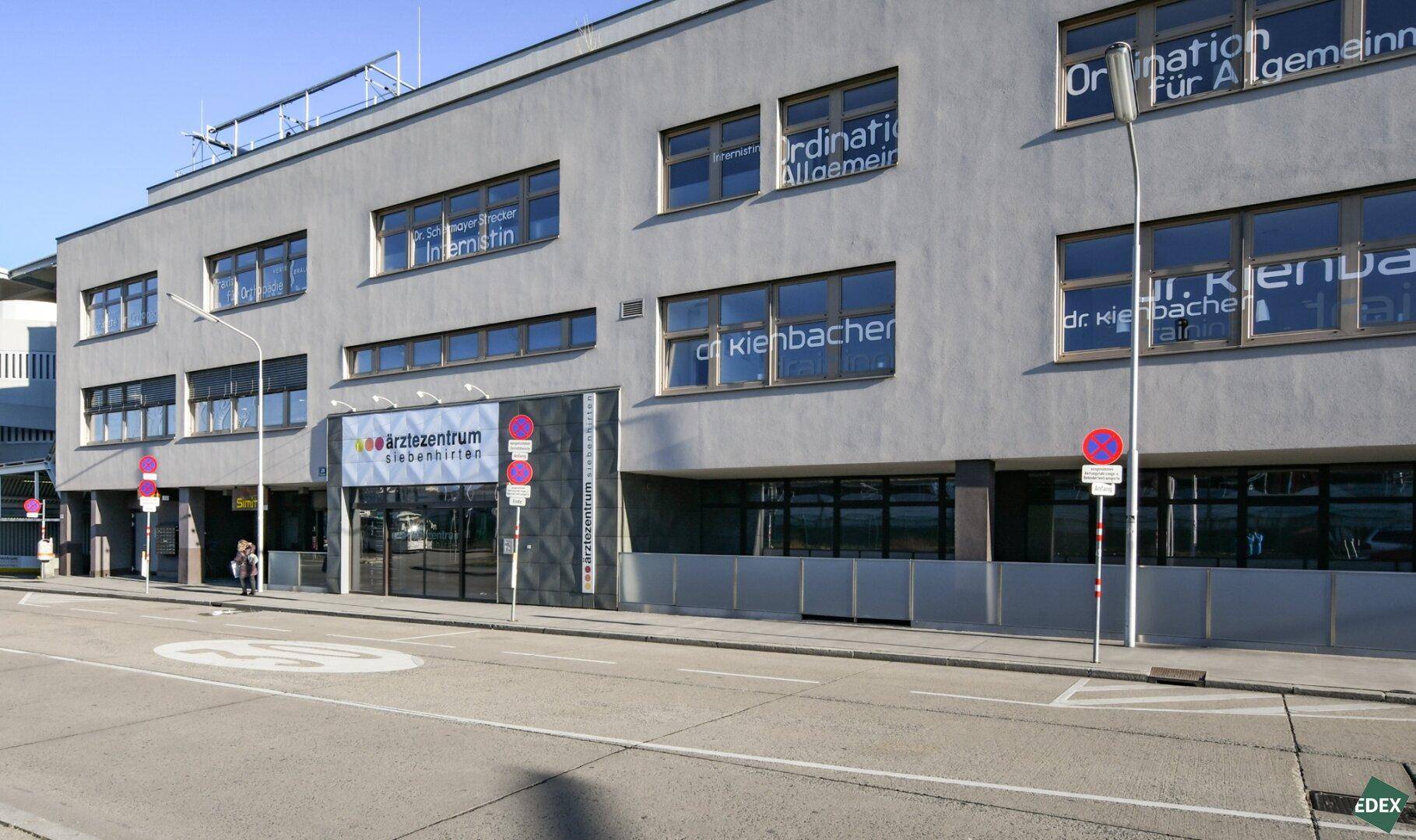 Weitwinkelansicht des modernen Ärztezentrums von der Straße aus, mit grauer Fassade und vielen Fenstern.