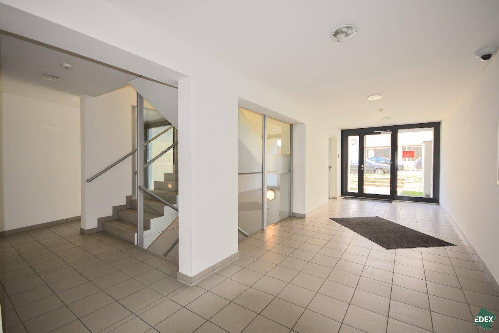 Heller Eingangsbereich des Gebäudes mit Treppenhaus, Fliesenboden und Glastüren.