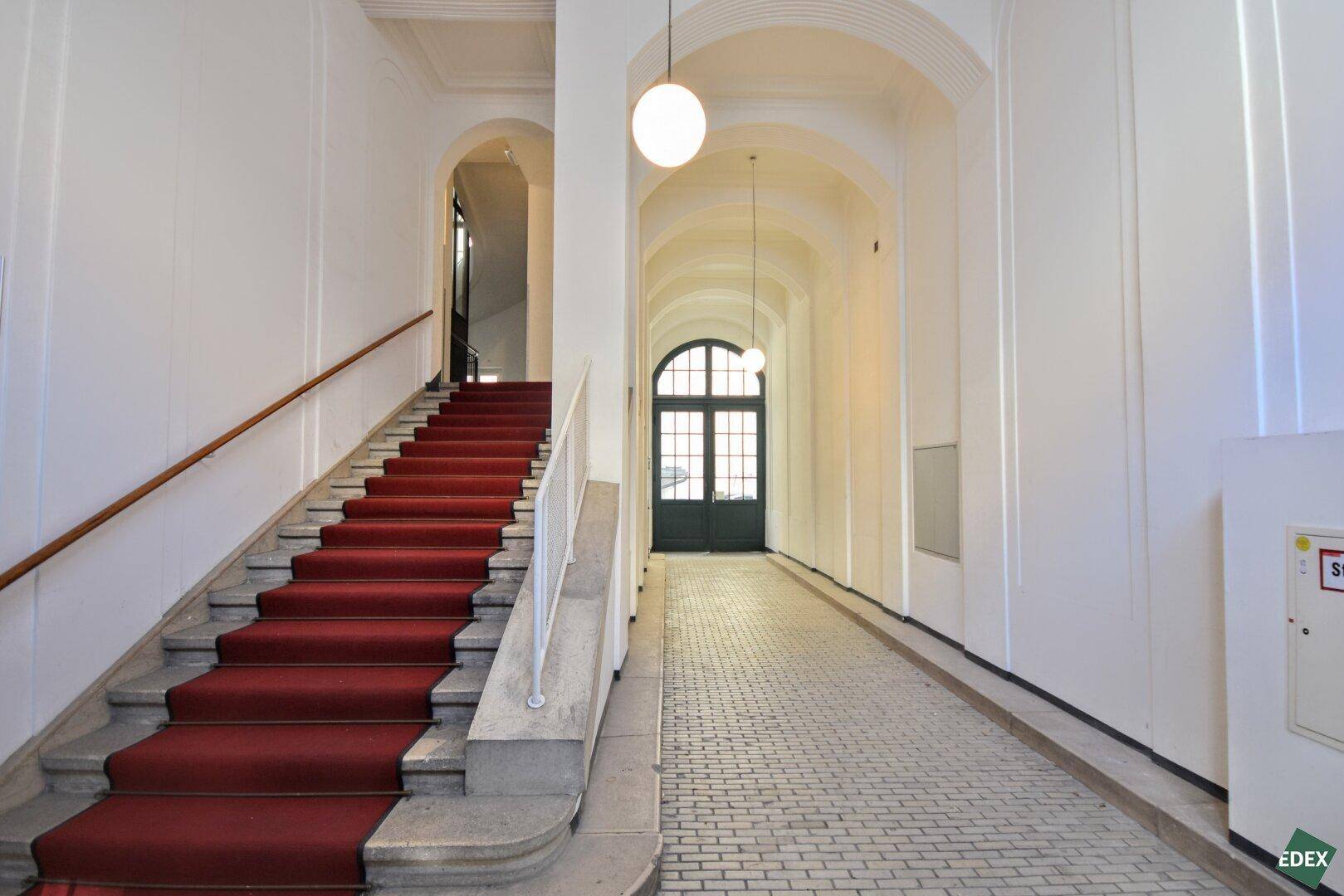 Elegantes Treppenhaus mit rotem Teppich, Marmorstufen und hohen Gewölbedecken, die zu einem großen Eingang führen.