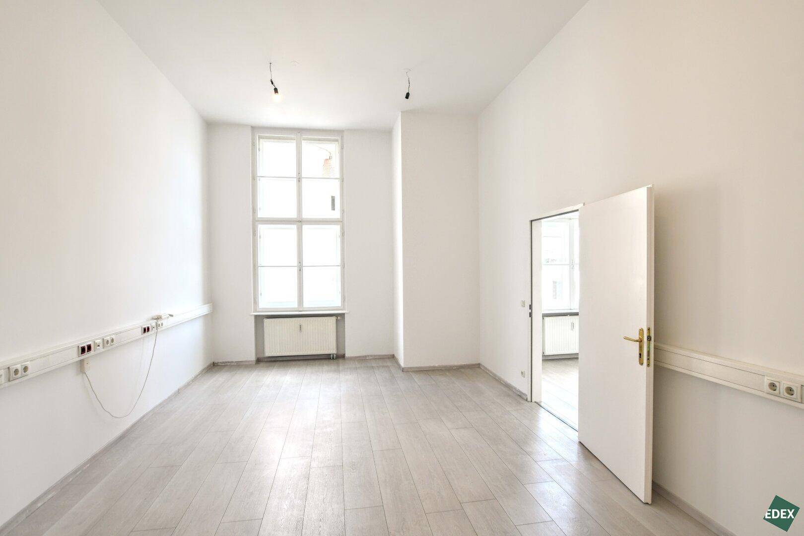 Kleinerer, heller Büroraum mit einem großen Fenster, hellem Holzboden und einer Tür zu einem Nebenraum.