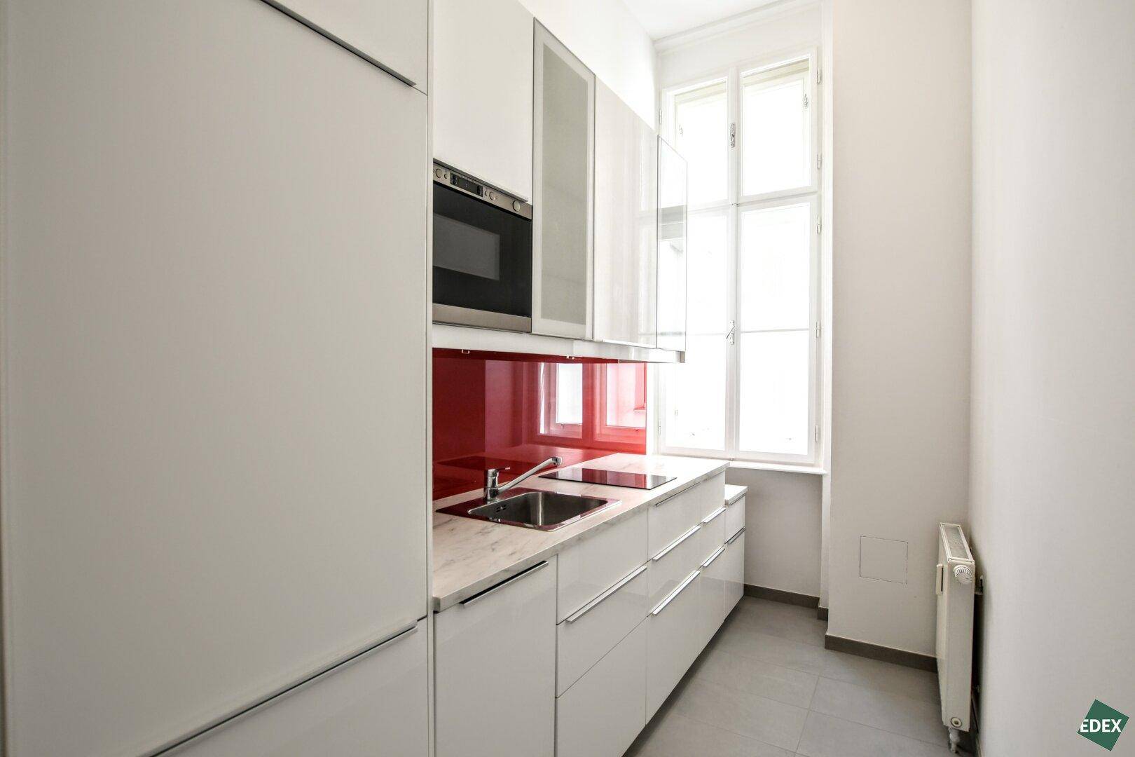 Moderne, schmale Küche mit weißen Schränken, roter Rückwand, Einbaugeräten und einem Fenster.