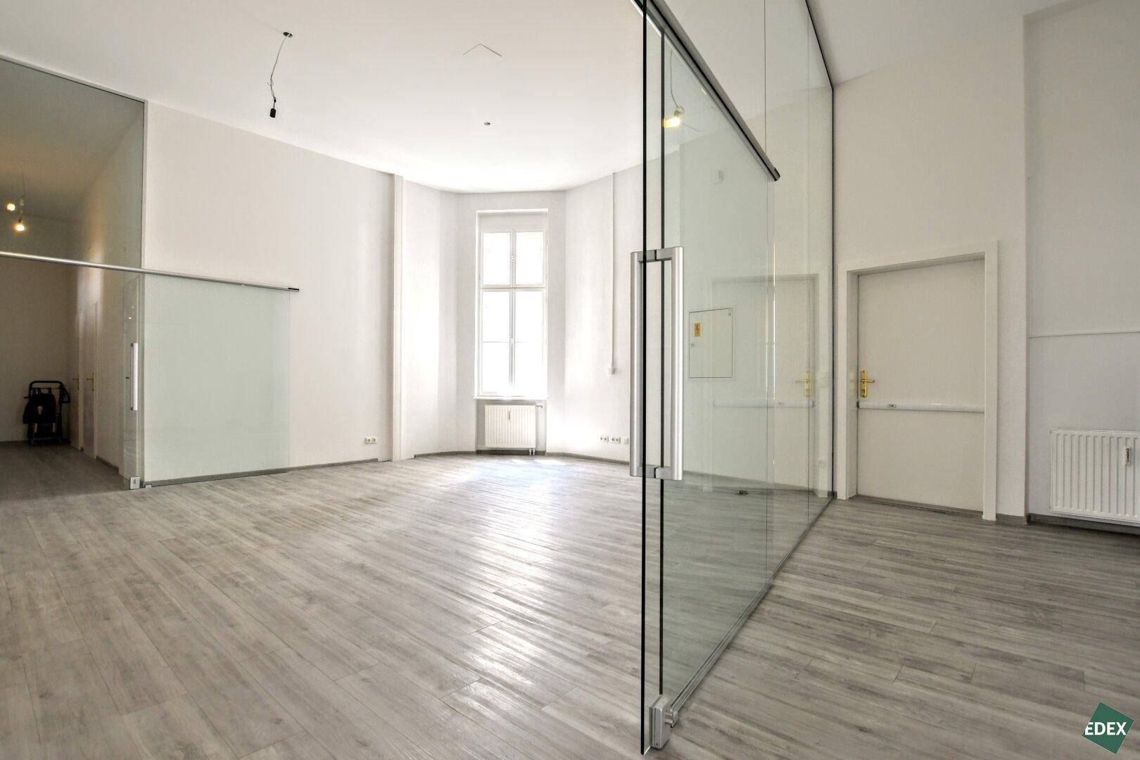 Heller Büroraum mit Holzboden, hohen Decken und einer eleganten Glastür, die zu einem weiteren Bereich führt.