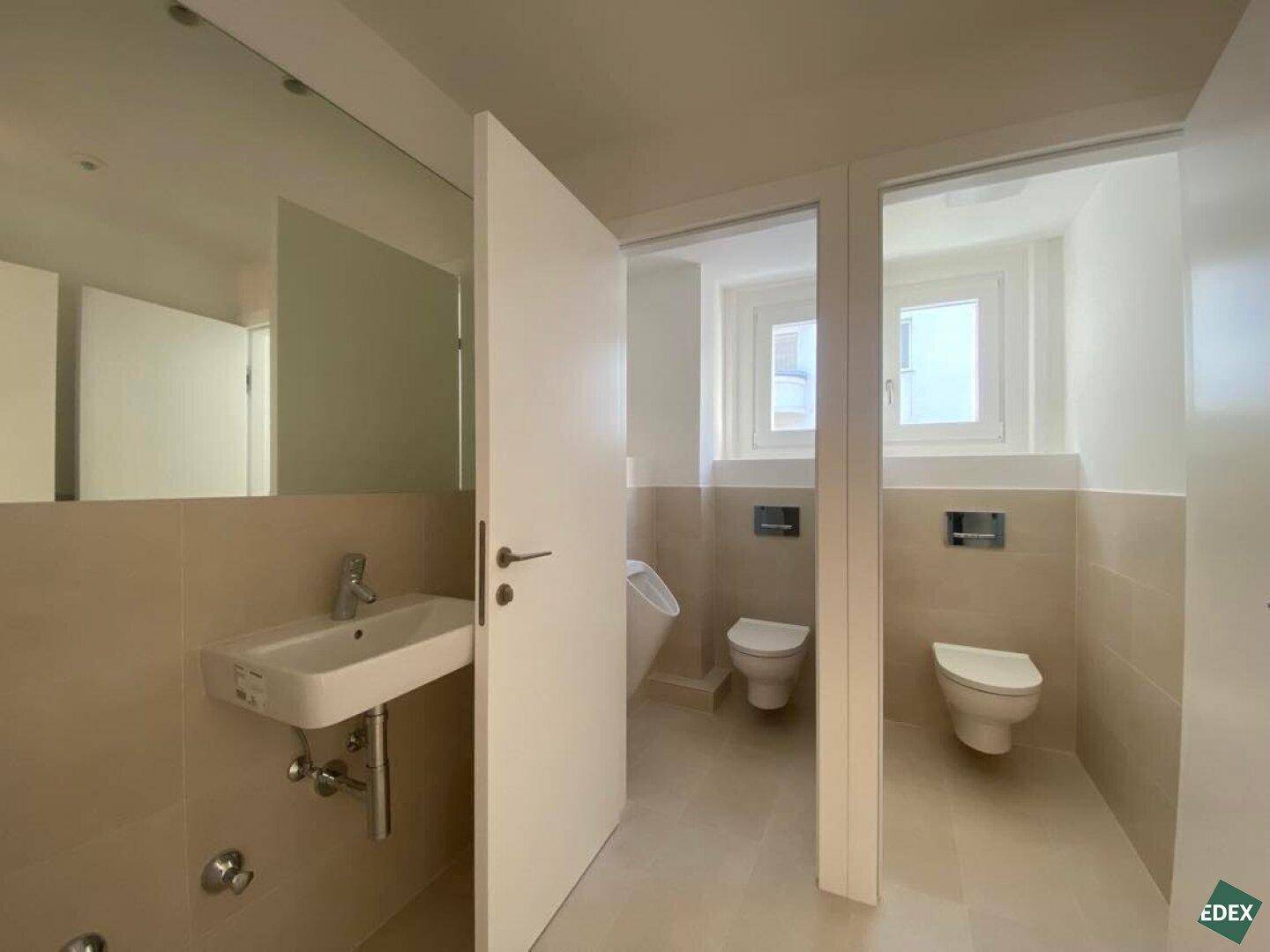 Modernes Badezimmer mit zwei Toiletten, Urinal und Waschbecken, ausgestattet mit beigen Fliesen.