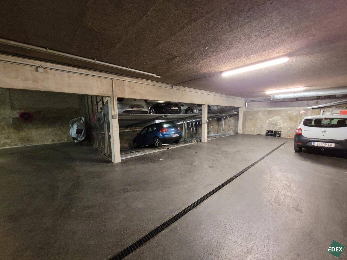 Weitwinkelansicht der Tiefgarage mit Parklifts, geparkten Fahrzeugen und einem gut beleuchteten Bereich.