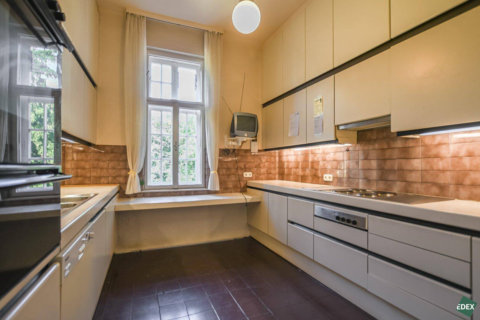 Küche mit älteren Einbauschränken, gefliestem Spritzschutz und Fenster mit Blick ins Grüne.