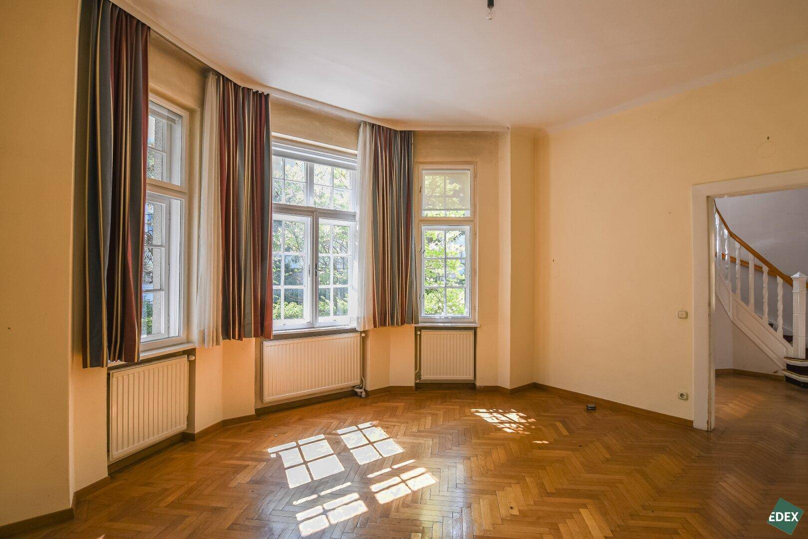 Helles Wohnzimmer mit Fischgrätparkett, großen Fenstern und viel Tageslicht.