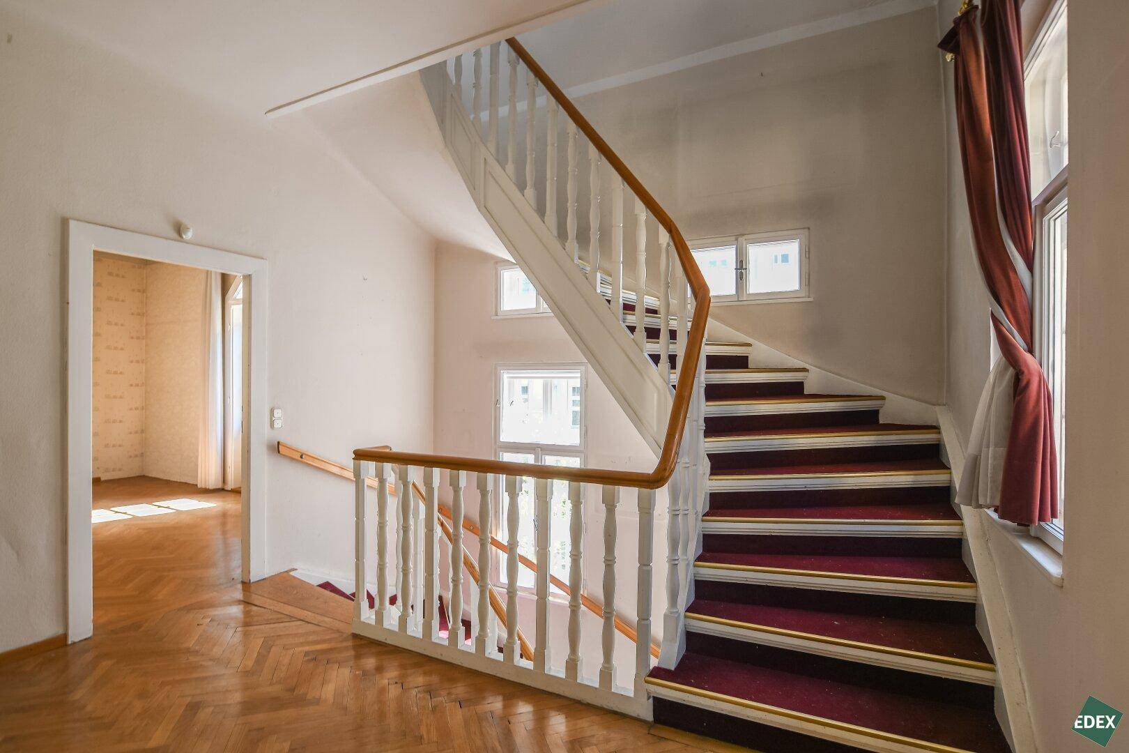 Helles Treppenhaus mit Teppichstufen, Holzgeländer und Fenstern für natürliches Licht.
