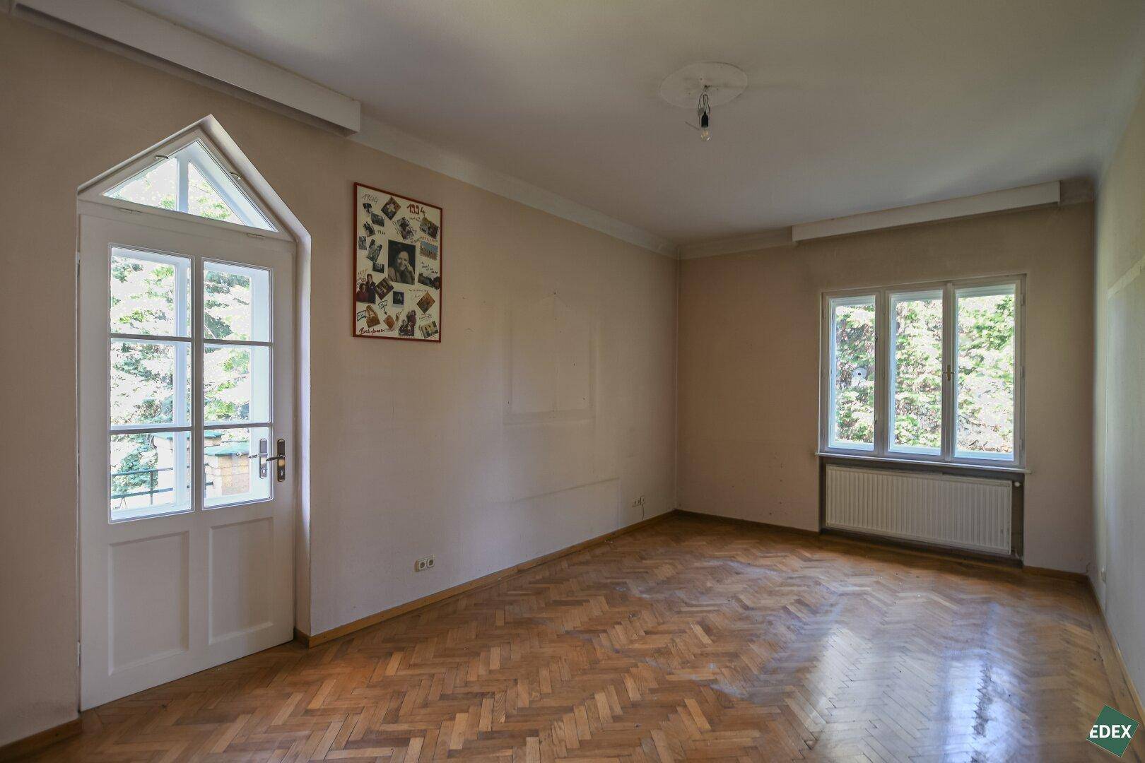 Leeres Zimmer mit Fischgrätparkett, Zugang zu einem Außenbereich und großem Fenster.