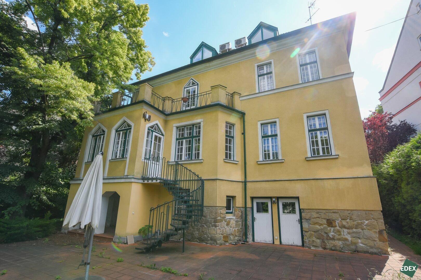 Historische gelbe Villa mit markanter Außentreppe und Steinsockel in sonniger Umgebung.