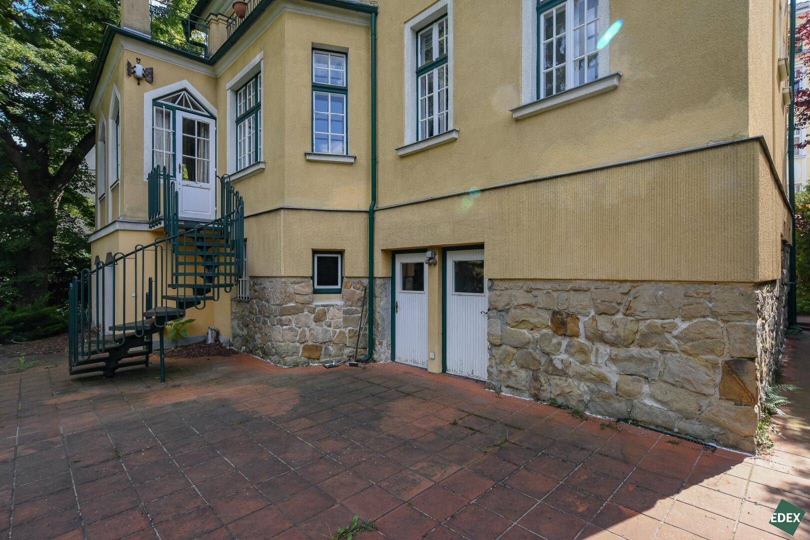 Seitenansicht der gelben Villa mit gepflastertem Bereich und eleganter Wendeltreppe.