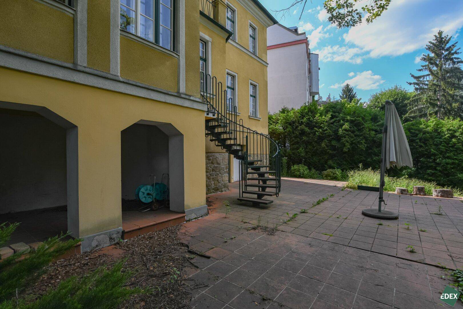 Gepflasterter Außenbereich neben der Villa mit Wendeltreppe und hoher Hecke.