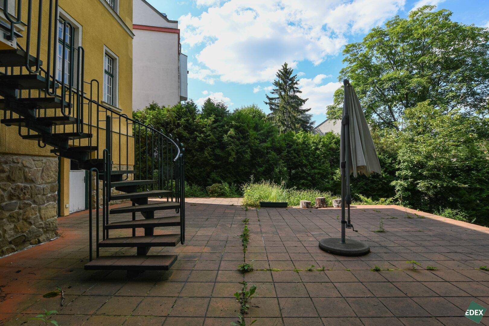 Gepflasterter Außenbereich mit Sonnenschirm, Wendeltreppe und Blick auf umliegende Gebäude.