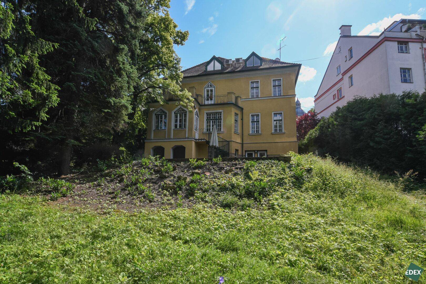 Gesamtansicht der gelben Villa von einem grünen Hang aus, umgeben von Bäumen.