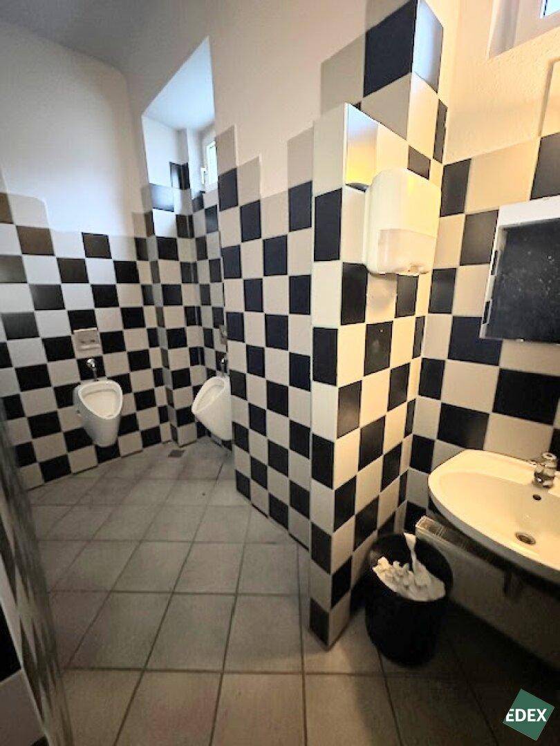 Öffentliche Toilette mit schwarz-weiß karierten Fliesen an den Wänden und Urinalen.