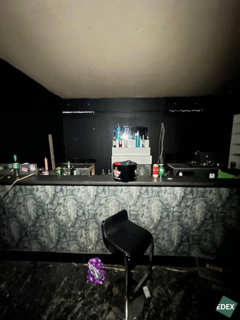 Bar-Bereich mit einer Theke, Barhockern und Flaschenregalen in einem dunklen Raum.