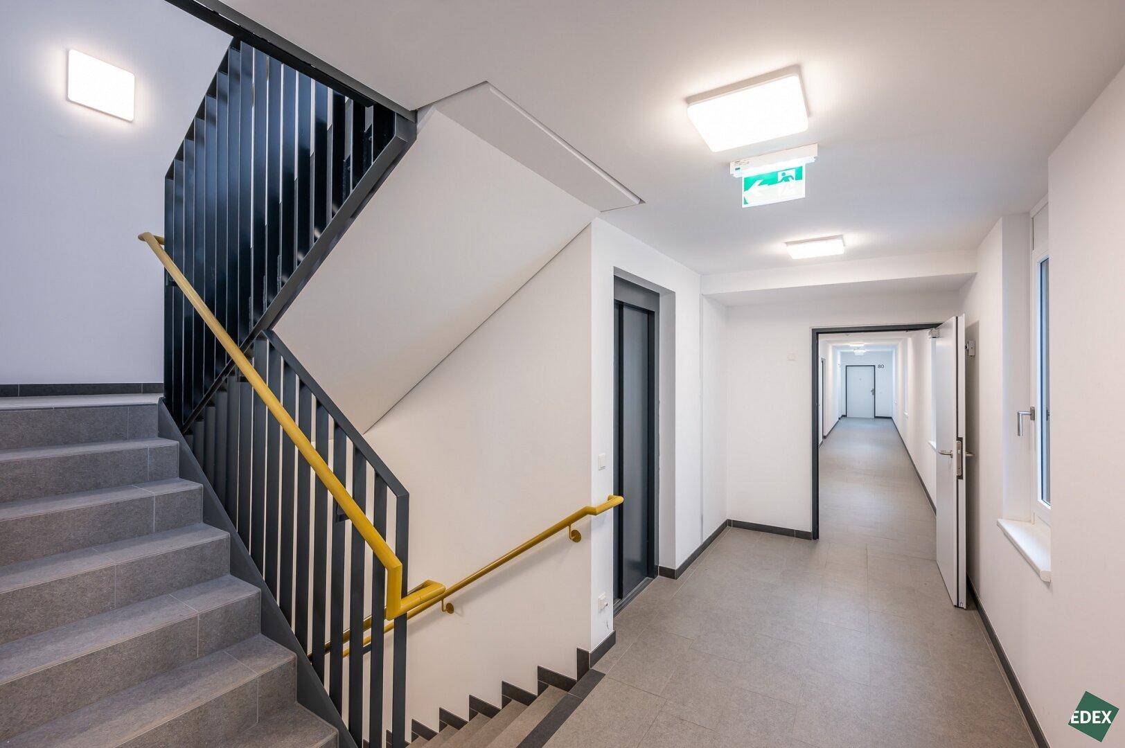 Modernes Treppenhaus mit grauem Fliesenboden und gelbem Handlauf, inklusive Aufzugzugang.