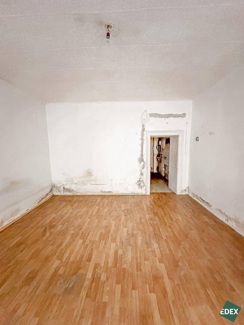 Leerer Raum mit Holzboden und einer Türöffnung, die in einen weiteren Bereich führt, mit sichtbaren Abnutzungsspuren.