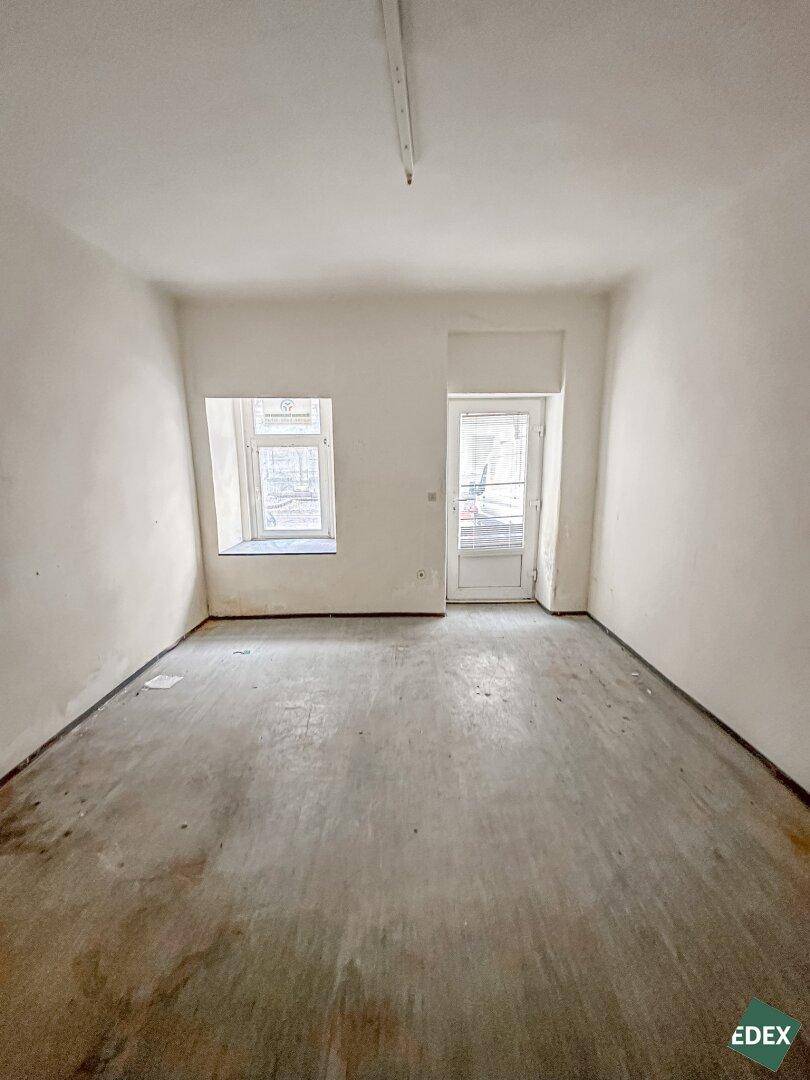 Leerer Raum mit grauem Betonboden, einem Fenster und einer Tür, die nach draußen führt, mit Renovierungsbedarf.