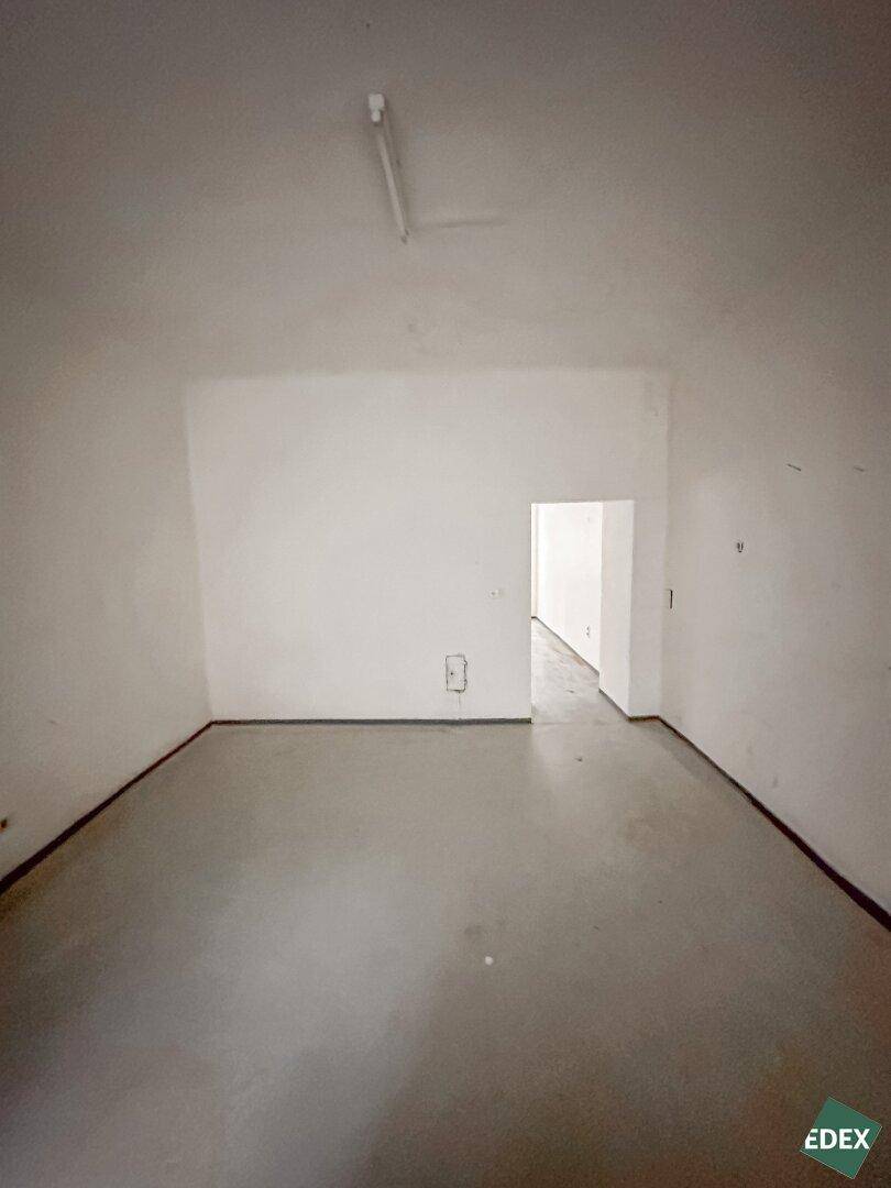 Leerer Raum mit grauem Betonboden und einer Türöffnung, die in einen helleren Bereich führt, mit einfacher Deckenbeleuchtung.