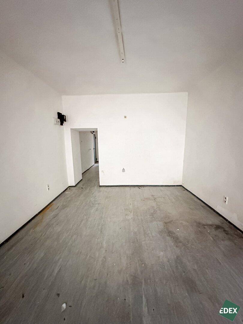 Leerer Raum mit grauem Betonboden und einem Durchgang zu einem Flur, der eine einfache Deckenbeleuchtung aufweist.