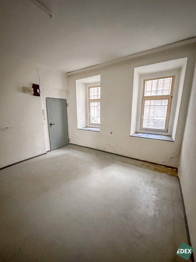 Leerer Raum mit grauem Betonboden, zwei Fenstern mit Gitterstäben und einer grauen Tür, die in einen anderen Bereich führt.