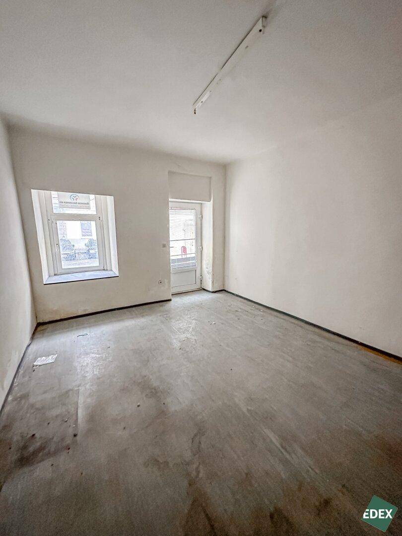 Leerer Raum mit grauem Betonboden, einem Fenster und einer Tür, die nach draußen führt, mit einfacher Deckenbeleuchtung.