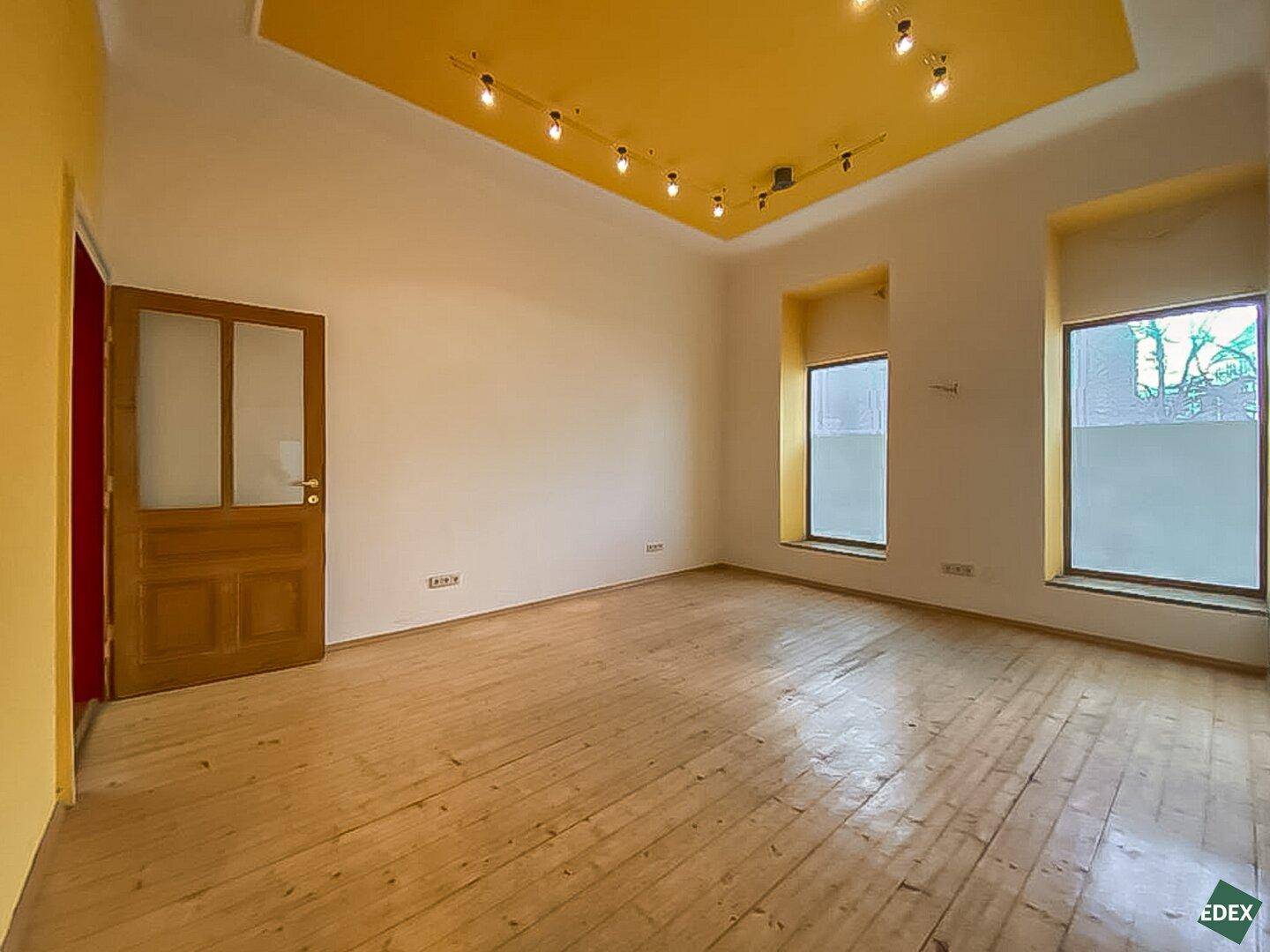 Heller Gewerberaum mit Holzdielenboden, gelber Decke und großen Fenstern, die viel Tageslicht hereinlassen.