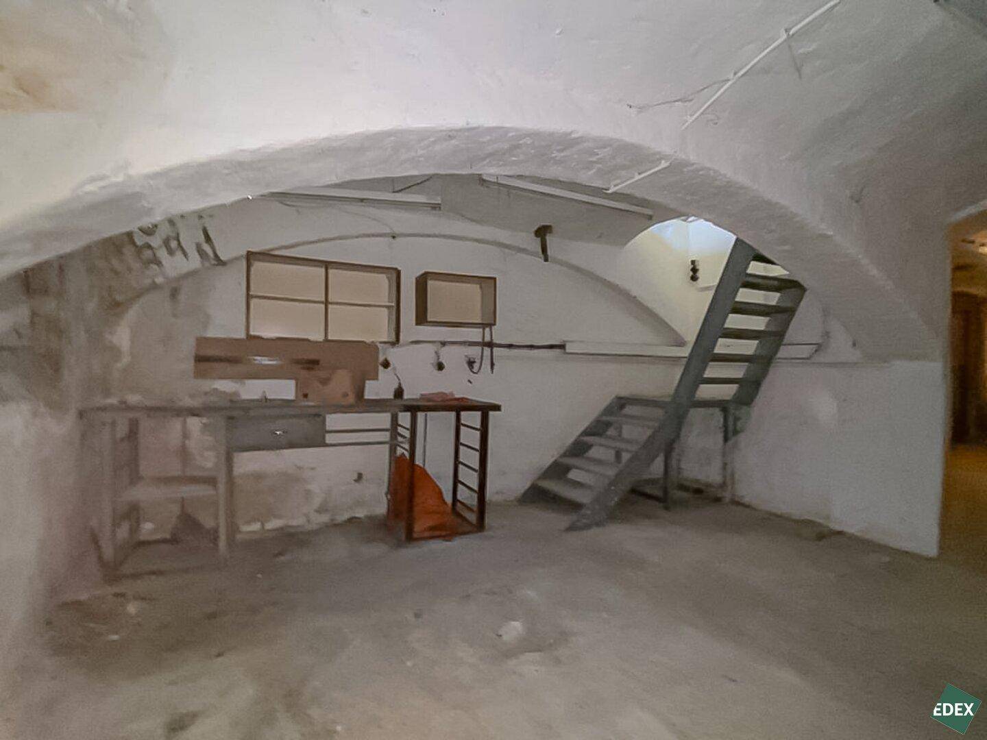 Unfertiger Gewölbekeller mit rohen Wänden, einer einfachen Holztreppe und sichtbaren Leitungen, in sanierungsbedürftigem Zustand.