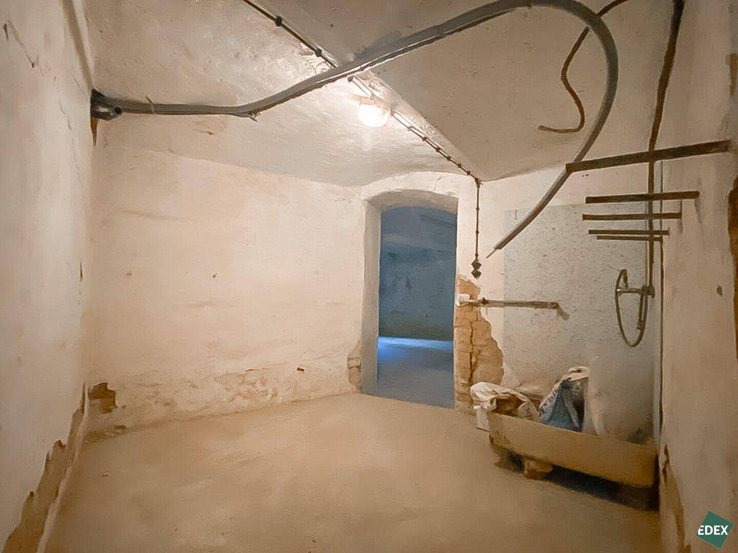 Unfertiger Kellerraum mit rauen Wänden, sichtbaren Leitungen und einem Durchgang zu einem weiteren Bereich, der auf Sanierung wartet.