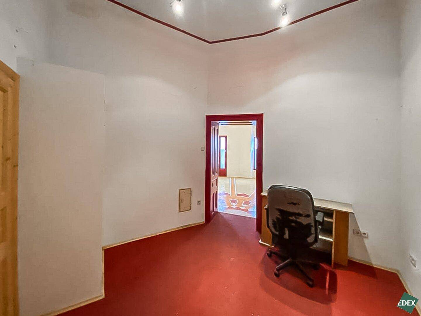 Kleiner Büroraum mit rotem Boden, weiß gestrichenen Wänden und einem Schreibtisch mit Bürostuhl, ideal für Einzelarbeit.