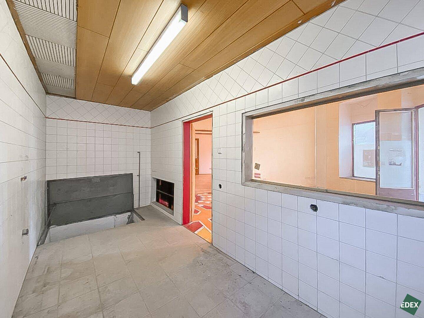 Gefliester Funktionsraum mit Holzdecke, einer Durchreiche und Blick in einen angrenzenden Bereich mit gemustertem Boden.