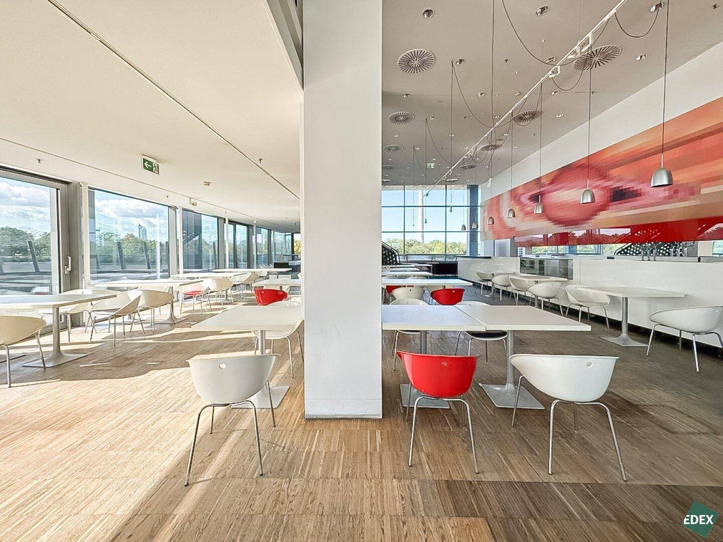 Moderne Cafeteria mit Holzboden, weißen Tischen und farbigen Stühlen, die einen hellen und einladenden Essbereich bietet.