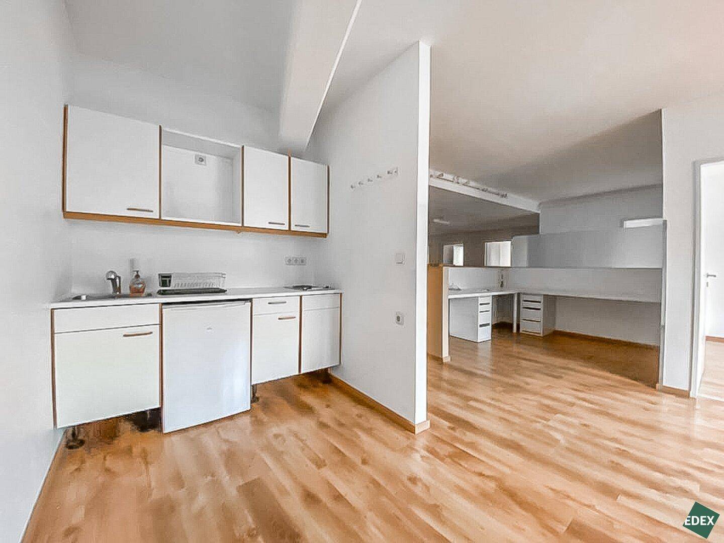 Kleine Küchenzeile mit weißen Schränken, Spüle und Kühlschrank, angrenzend an einen offenen Bürobereich.