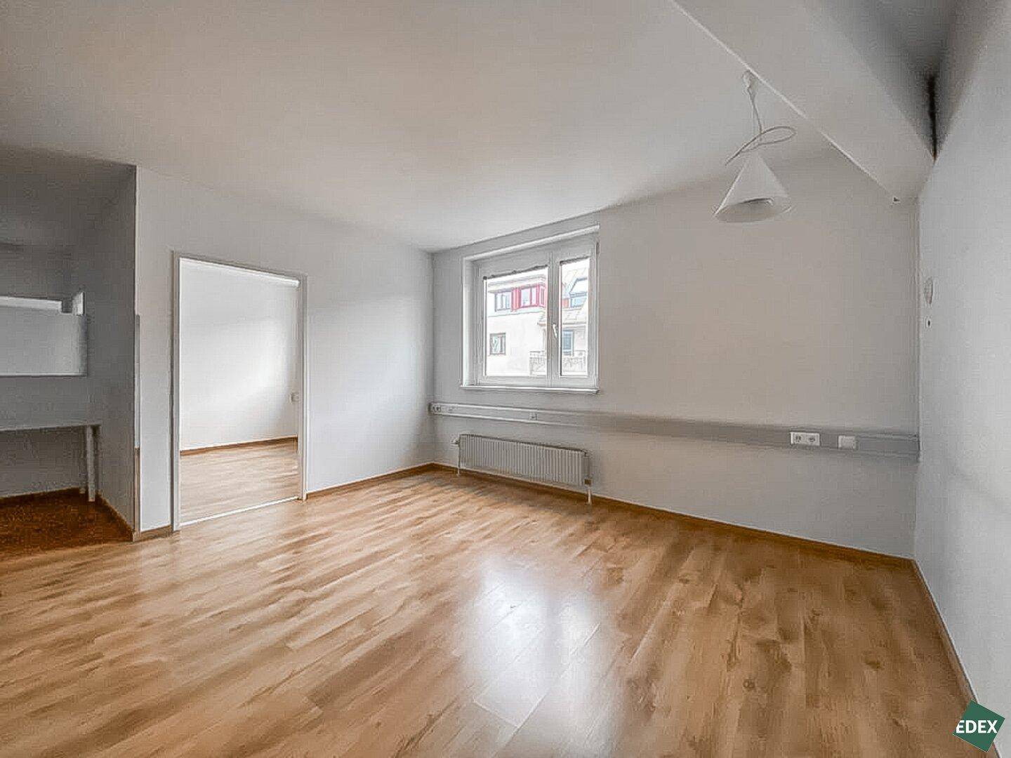 Helles Büro mit Holzfußboden, großem Fenster und Blick in einen weiteren Raum.