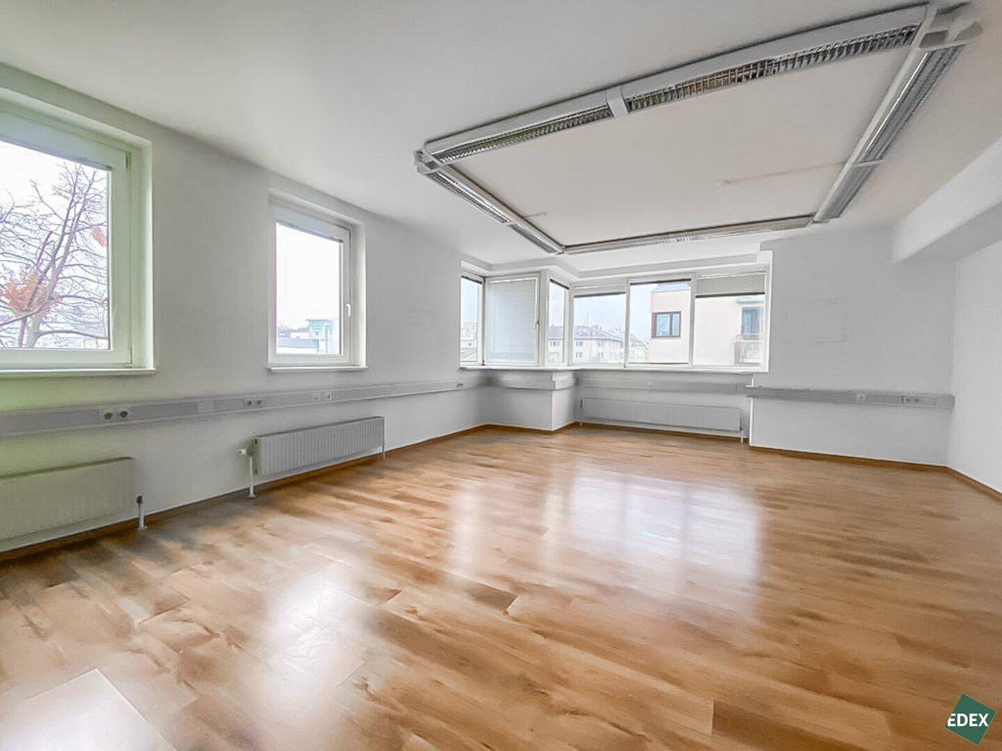 Helles Büro mit großen Fenstern und Holzfußboden, ausgestattet mit Deckenleuchten und Heizkörpern.
