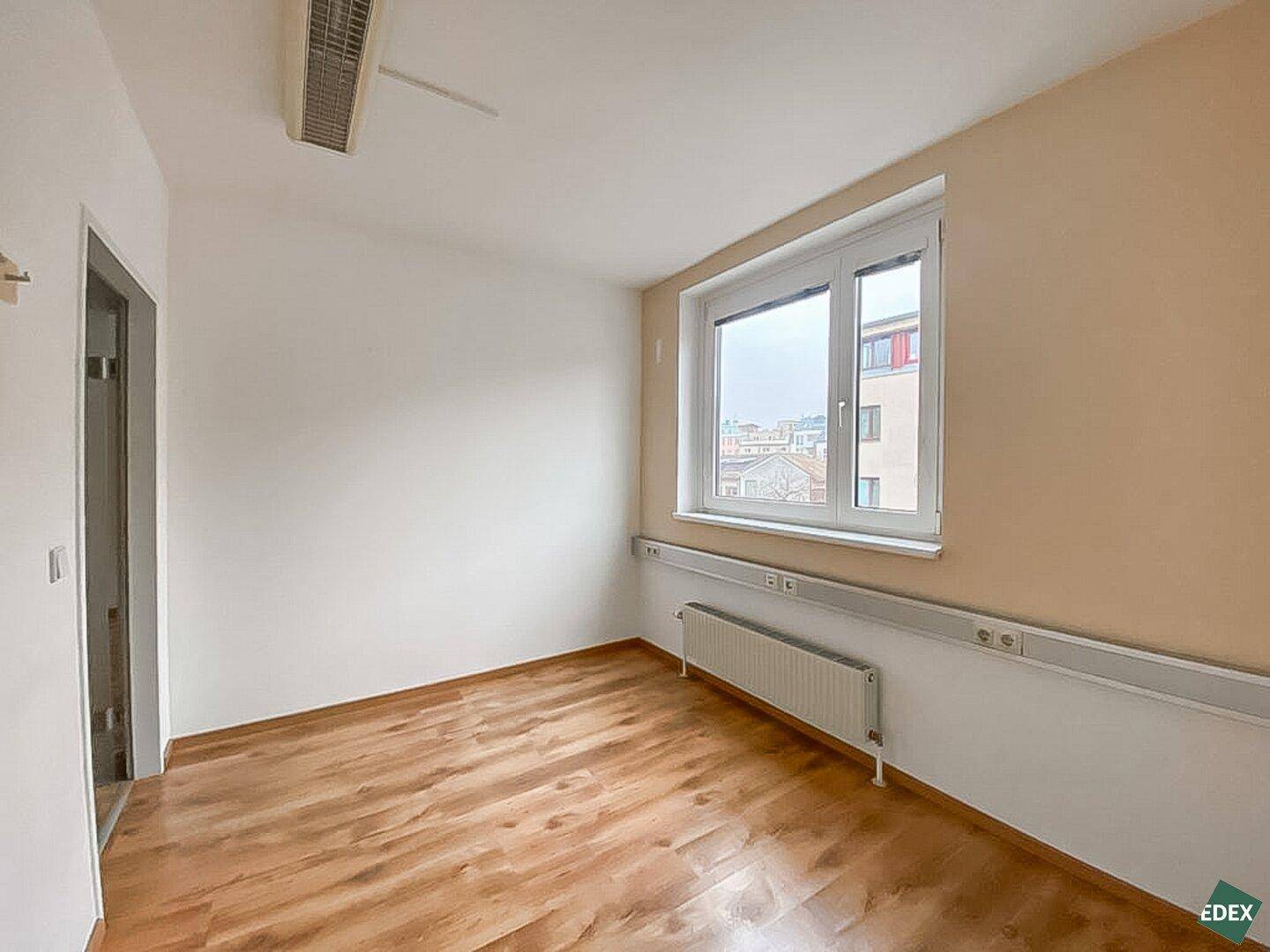 Büro mit Holzfußboden, großem Fenster und einer beigen Akzentwand, gut beleuchtet.