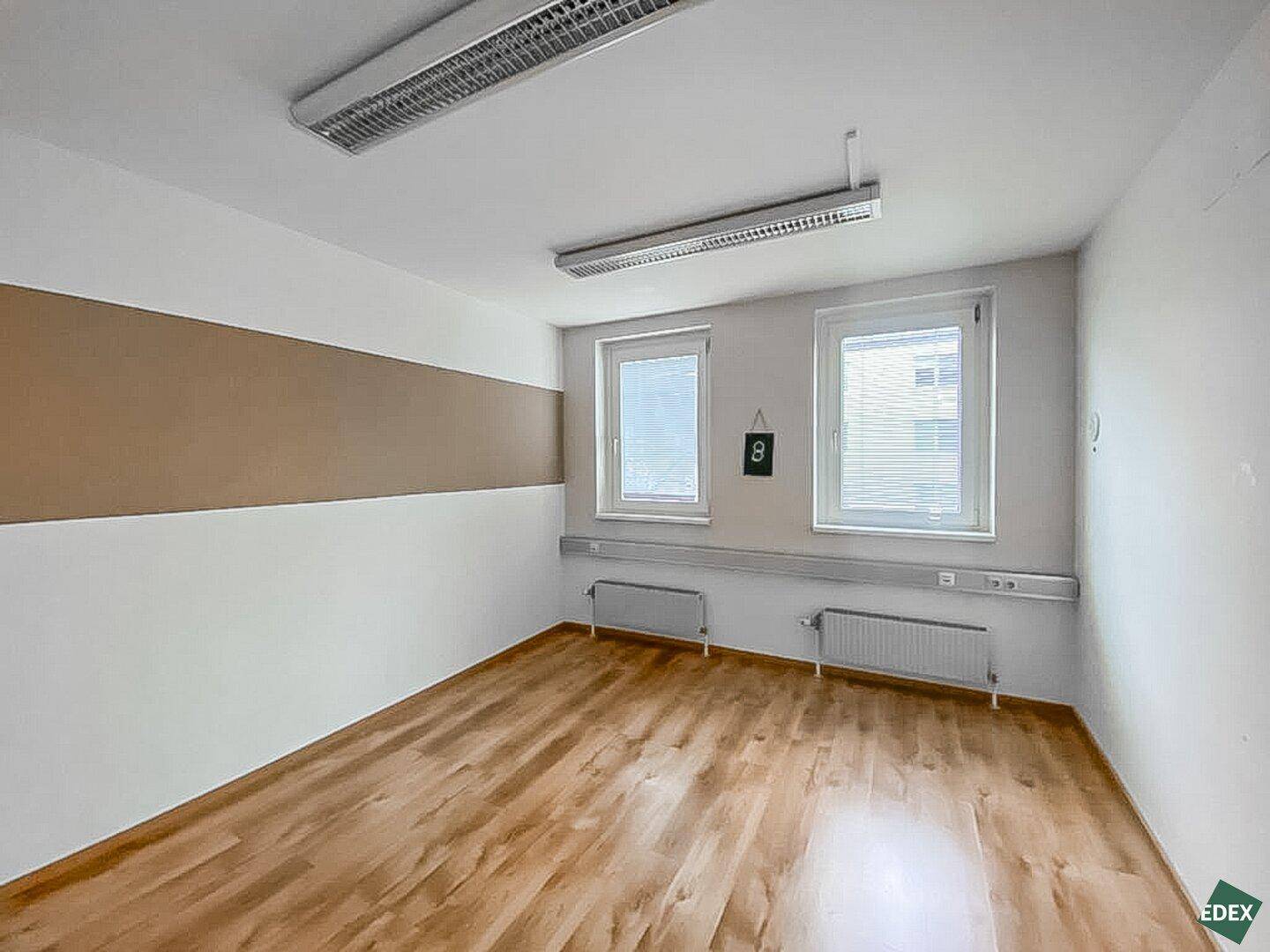 Büro mit Holzfußboden, zwei Fenstern und einer beigen Akzentwand, gut beleuchtet.