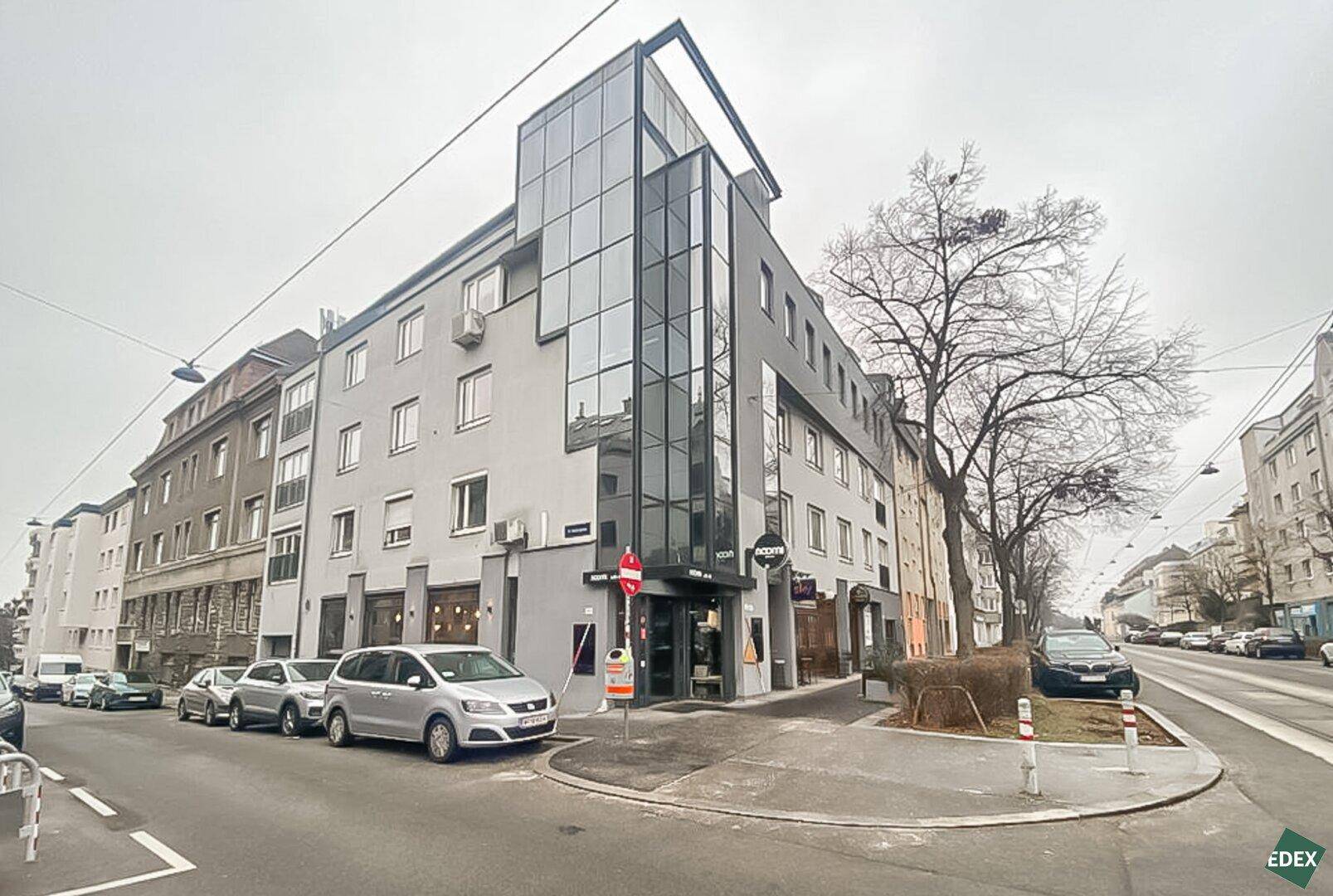 Modernes Eckgebäude mit Glasfassade und mehreren Etagen, umgeben von städtischer Bebauung.